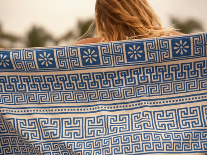 Close-up detail of the Mollusk Surf Shop Poseidon Towel held up outdoors, showing blue and natural white jacquard weave with Greek key meander pattern and daisy floral motifs