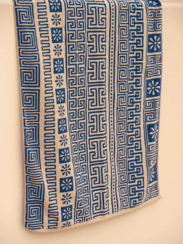 Mollusk Surf Shop Poseidon Towel draped over a towel bar, showing full blue and white jacquard pattern with Greek key meander, daisy motifs, and striped border detail