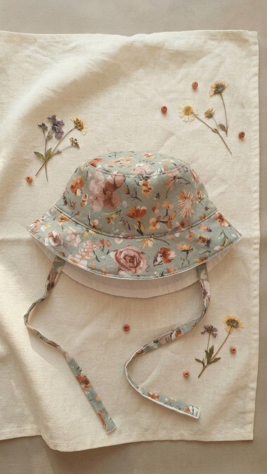 A flat lay photograph of the Primavera baby bucket hat in a soft sage green fabric with a white paisley and floral pattern. The hat features a matching chin strap and a small green turtle brand label on the brim. It is styled on a neutral linen surface next to a sprig of dried lavender and a small stone in soft, natural light.