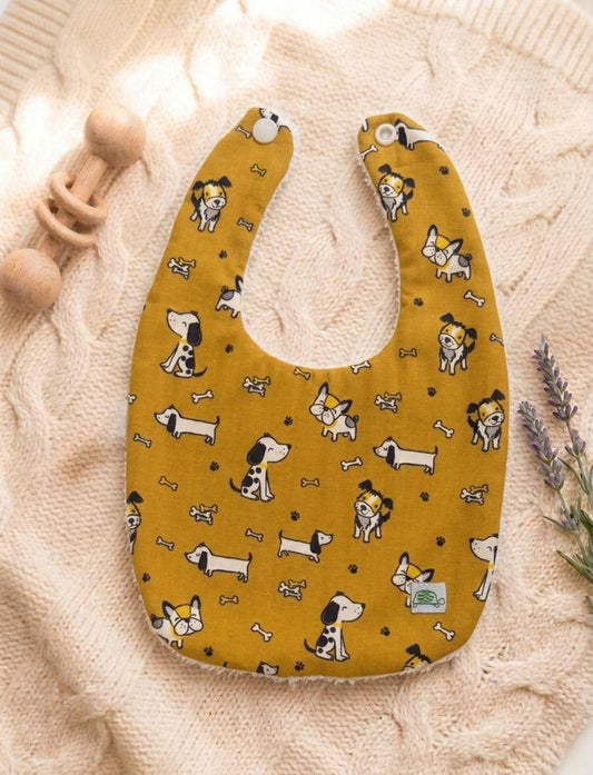 A flat lay of a mustard yellow baby bib featuring a whimsical pattern of various cartoon puppies, tiny bones, and paw prints. The bib has white snap closures at the top and is resting on a soft pink cable-knit sweater alongside a wooden rattle and lavender sprigs.