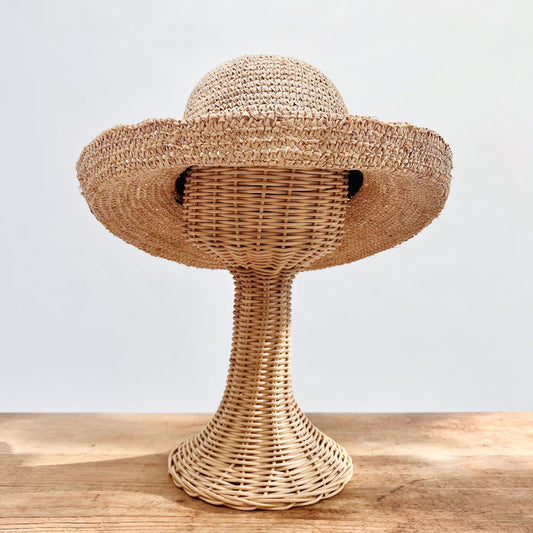 Natural raffia kettle brim hat with wide floppy brim and open block crochet weave, shown on a wicker hat stand — front view