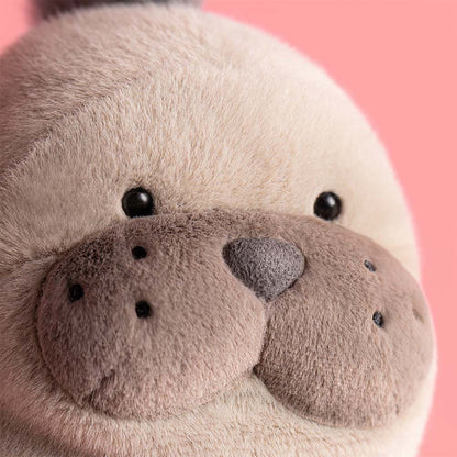 Close-up of Jellycat Roarwell Sea Lion's face showing soft cream fur, large rounded brown muzzle cheeks with embroidered dots, a grey nose, and black bead eyes, against a pink background