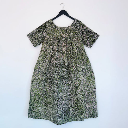 Full front view of the Sakina Dress in Green and Pink Speckle, hanging on a black hanger against a white wall, showing a round neckline, short sleeves, gathered yoke, voluminous A-line silhouette, and hand-printed green, pink, and white speckle pattern on 100% cotton Batiki fabric
