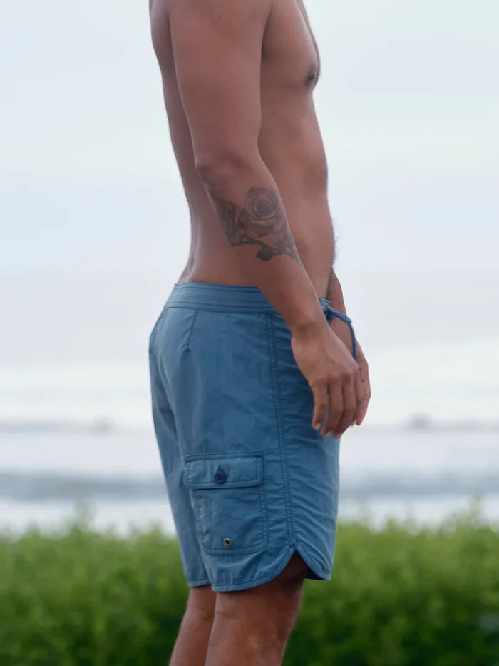 Male model wearing the Mollusk Surf Shop Scallop Trunks in Indigo, side profile showing indigo blue boardshorts with scallop curved hem, back cargo pocket with snap closure, photographed outdoors with ocean in background