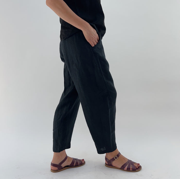 Person with their hands in their pockets wearing black linen pants and a black linen tank on a white background as seen from the side.