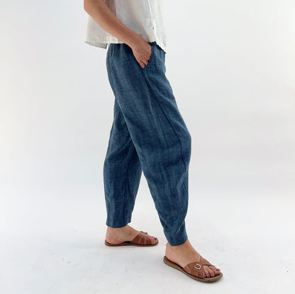 Person wearing blue melange linen pants and a white top and brown sandals, with their hands in their pockets on a white background.
