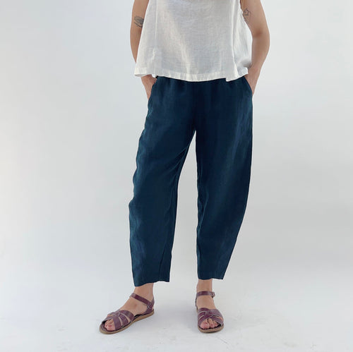 Person wearing navy linen pants and a white linen top on a plain background. Their hands are in their pockets.