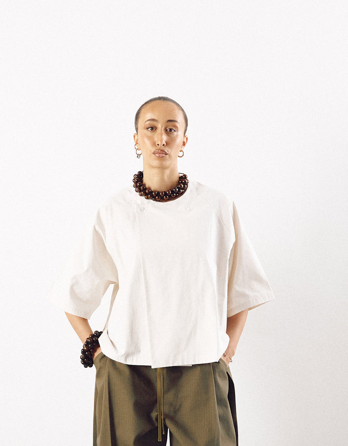 Model wearing an off-white minimalist short sleeve shirt with a boxy fit, paired with olive pants and accessorized with chunky dark bead jewelry, highlighting understated elegance.