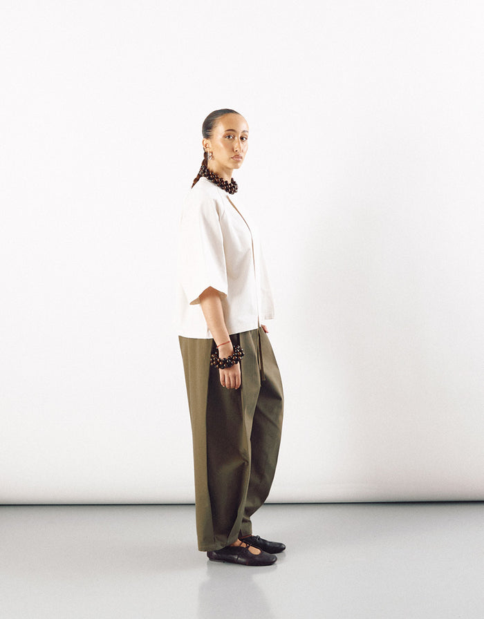 Side view of a model wearing an off-white short sleeve minimalist shirt paired with wide-leg olive green pants, styled with dark accessories and black shoes.