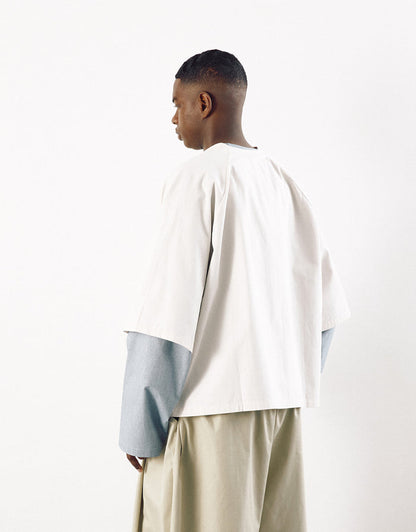 Man wearing a minimalist off-white short sleeve shirt layered over a long-sleeve garment, paired with loose beige trousers, showcasing contemporary design and understated elegance.