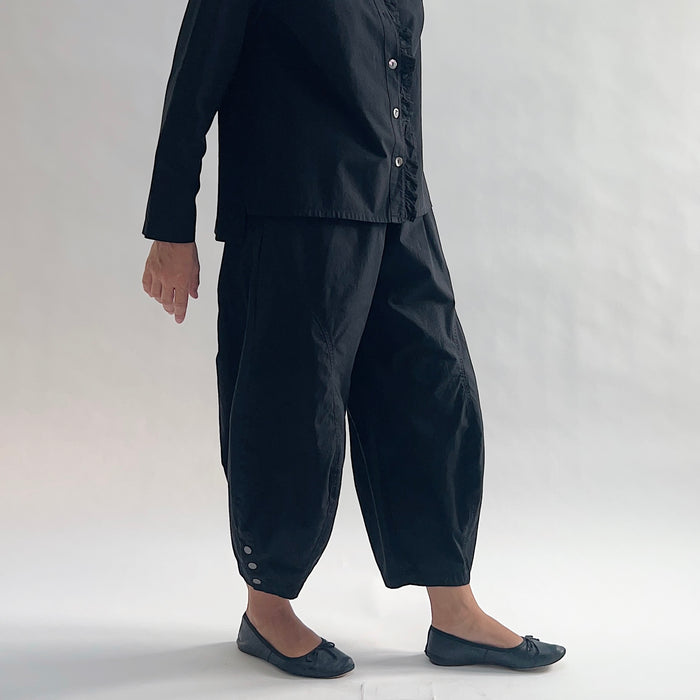 Side view of model with hand on hip wearing Eleven Stitch black snap doreen pant, showing elastic waistband, wide balloon leg tapering to snap hem at ankle, side seam pocket, snap buttons at ankle, and black color, styled with matching black blossom collar shirt and black ballet flats