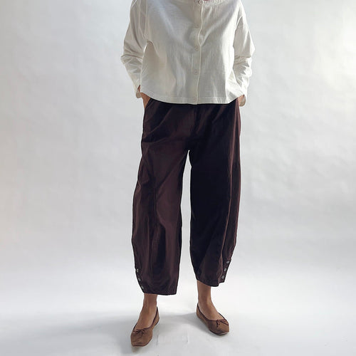 Full front view of model wearing Eleven Stitch chicory snap doreen pant with hands in pockets, showing elastic waistband, wide balloon leg tapering to snap hem at ankle, and deep espresso brown color, styled with cream button front shirt and taupe ballet flats