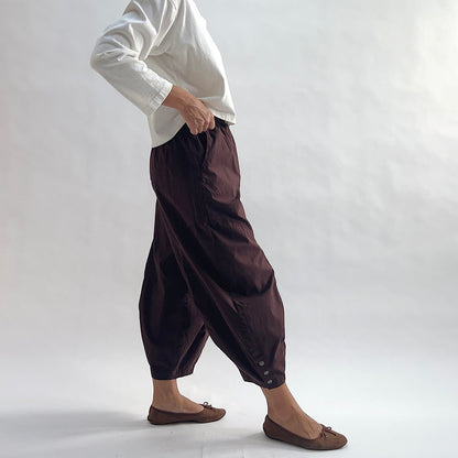 Side view of model wearing Eleven Stitch chicory snap doreen pant, showing elastic waistband, wide balloon leg tapering to snap hem at ankle, side seam pocket, and deep espresso brown color, styled with cream long sleeve top and taupe ballet flats