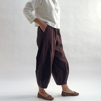 Three-quarter front view of model wearing Eleven Stitch chicory snap doreen pant with hand in pocket, showing elastic waistband, wide balloon leg tapering to snap hem at ankle, side seam pocket, and deep espresso brown color, styled with cream button front shirt and taupe ballet flats