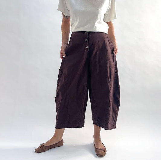 Full front view of model wearing Eleven Stitch chicory snap front pant, showing snap front waistband, wide balloon leg, large side patch pockets, gathered elastic ankle hem, and deep espresso brown color, styled with cream short sleeve tee and taupe ballet flats