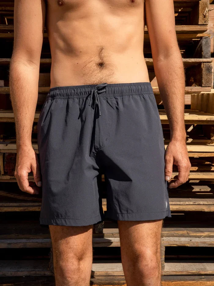 Male model wearing the Mollusk Surf Shop Stretch Vacation Trunks in Navy, front view showing navy nylon shorts with elastic waistband and drawstring, photographed outdoors against stacked wooden pallets in sunlight