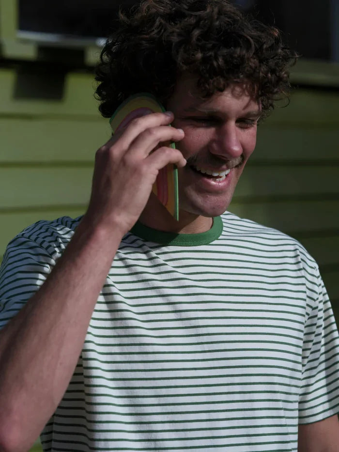 A smiling man with curly brown hair talking on a phone, wearing a white t-shirt with thin green horizontal stripes and a green collar, set against a green house siding background.