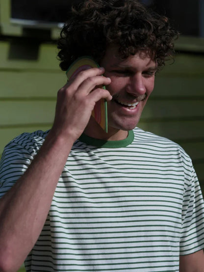 A smiling man with curly brown hair talking on a phone, wearing a white t-shirt with thin green horizontal stripes and a green collar, set against a green house siding background.