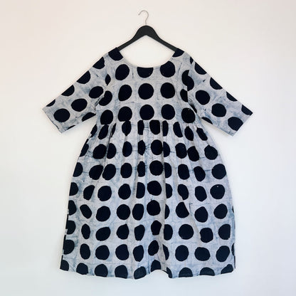 Full front view of the Sula Dress in Black Indigo Dots, hanging on a black hanger against a white wall, showing a round neckline, elbow-length sleeves, gathered waist seam, and oversized hand-printed black polka dots on grey-white 100% cotton Batiki fabric