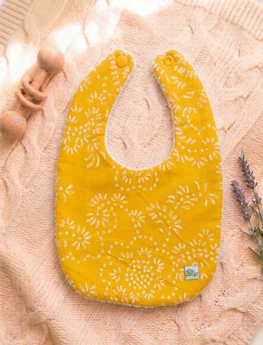 A flat lay photograph of a U-shaped baby bib made from mustard yellow muslin cotton. The bib features a delicate, etched white floral and scrollwork pattern across the front and has a coordinating yellow snap closure. It is resting on a pink textured cable-knit sweater, next to a wooden rattle and sprigs of lavender. A small logo tag with a turtle is visible on the bottom edge.