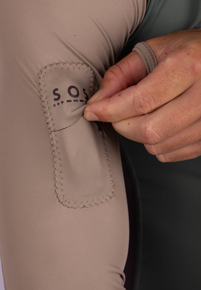 Close-up of the S.O.S.-branded side pocket on the Waihana Terra Hooded Spearfishing Rashguard, with a hand demonstrating the pocket opening against a tan and olive fabric body.