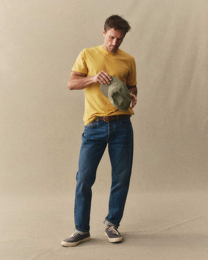 Man wearing Taylor Stitch Cotton Hemp Tee in Goldfinch yellow with blue jeans and sneakers, holding an olive hat