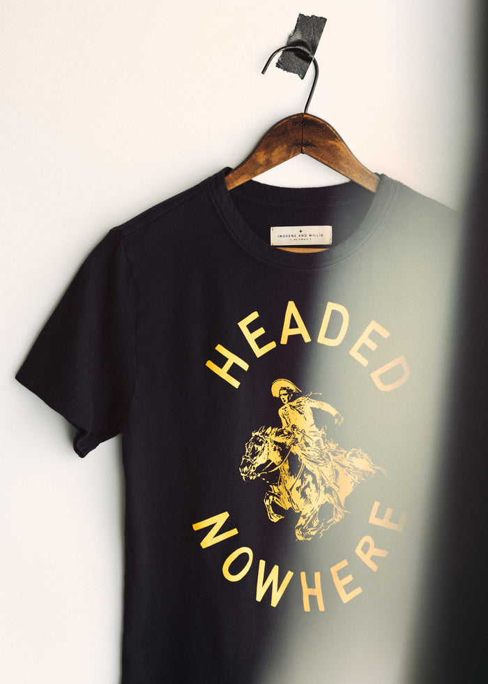 Angled close-up of The Headed Nowhere Tee by Imogene + Willie on a hanger, showing the Imogene and Willie label at the neckline and the gold screen-printed 'HEADED NOWHERE' circular graphic with cowboy on horseback on a black cotton tee