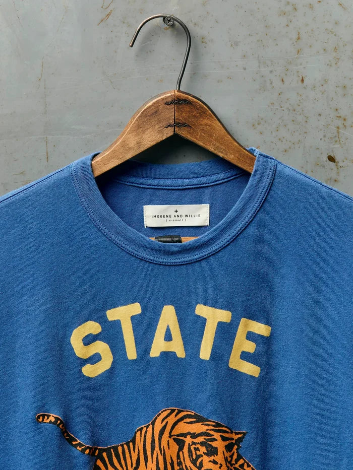 Close-up of the neckline of The State Tee in Blue showing the Imogene and Willie label, ribbed crew neck, and the top of the gold 'STATE' screen print with tiger graphic on blue cotton