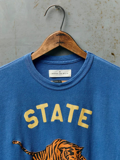 Close-up of the neckline of The State Tee in Blue showing the Imogene and Willie label, ribbed crew neck, and the top of the gold 'STATE' screen print with tiger graphic on blue cotton