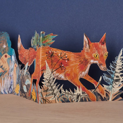 Close-up detail of the Three Fold Fox concertina card showing a vividly illustrated orange fox mid-stride through ferns and winter botanicals, with birds visible on the adjacent panel