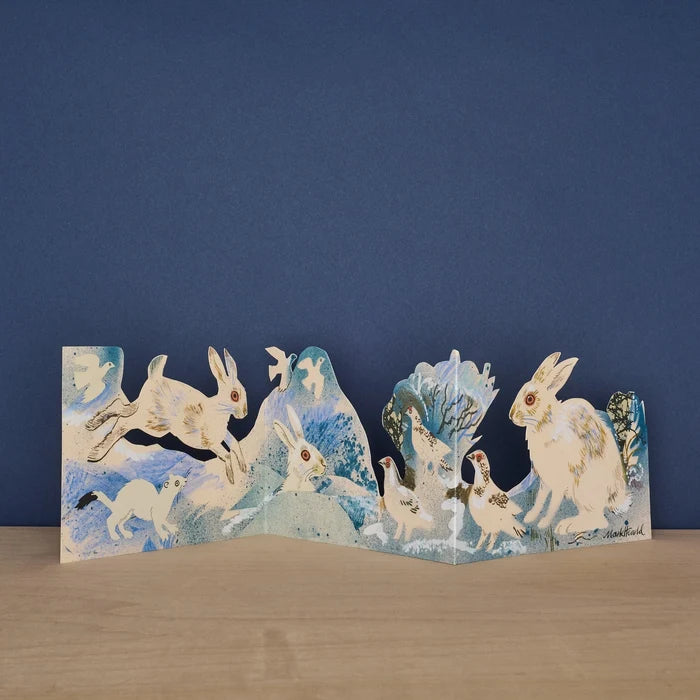 Three Fold Winter Wildlife concertina card by Mark Hearld, fully extended to show white hares leaping through a snowy landscape alongside a stoat and white doves, against a navy background