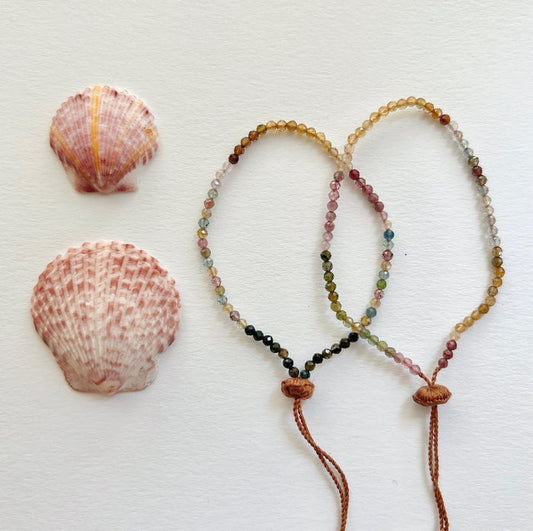 Two tourmaline tassel bracelets on a white surface, accompanied by two seashells. One bracelet is predominantly pink and green, and the other is multicolored with an adjustable tassel closure.