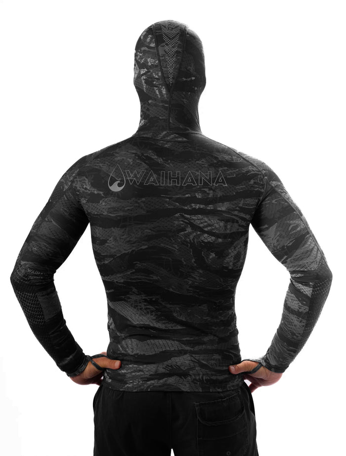 Back view of a male model wearing the Waihana Tropicam Black hooded rashguard in a dark tactical camouflage pattern with mesh panels on the shoulders and forearms, hands on hips against a white background.
