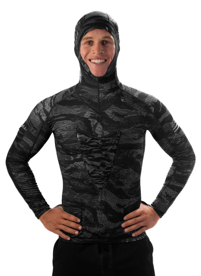 Male model wearing the Waihana Tropicam Black hooded rashguard in a dark tactical camo pattern, hands on hips, smiling against a white background.