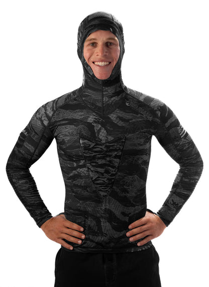 Male model wearing the Waihana Tropicam Black hooded rashguard in a dark tactical camo pattern, hands on hips, smiling against a white background.