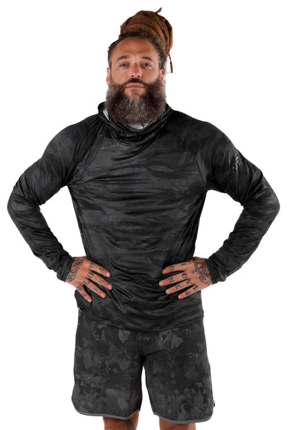 Male model wearing the Waihana Tropicam Black long-sleeve sun shirt with integrated neck gaiter in a dark camouflage pattern, paired with matching camo shorts, hands on hips against a white studio background.