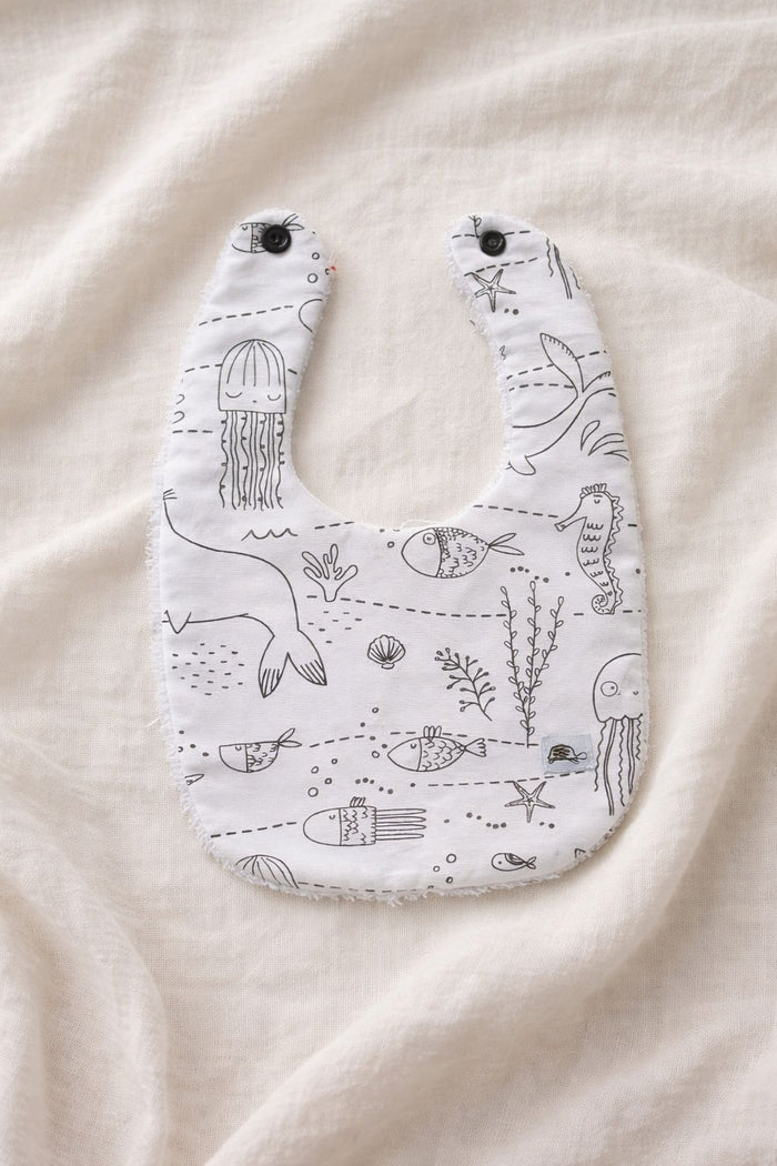 A flat lay photograph of a handmade baby bib featuring a white fabric with black line-art illustrations of undersea life, including jellyfish, seahorses, fish, and coral. The bib has two black snap fasteners at the neck and a soft white towelling texture visible along the edges. It is styled against a neutral, draped cream-colored fabric background.