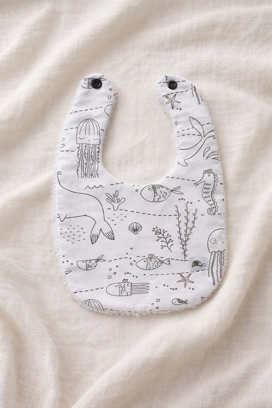 A flat lay photograph of a handmade baby bib featuring a white fabric with black line-art illustrations of undersea life, including jellyfish, seahorses, fish, and coral. The bib has two black snap fasteners at the neck and a soft white towelling texture visible along the edges. It is styled against a neutral, draped cream-colored fabric background.