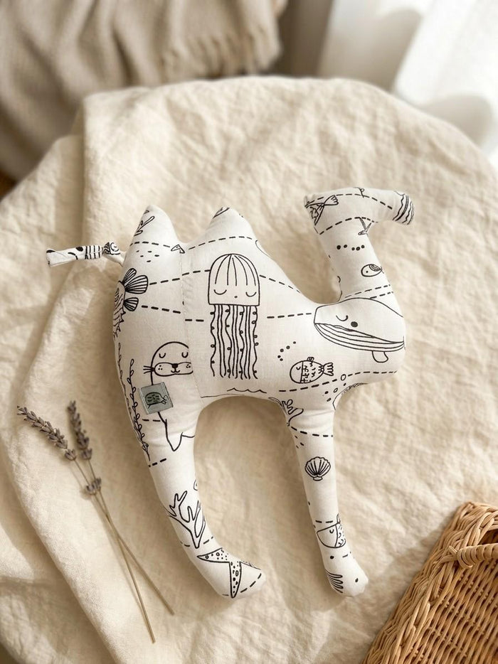 A handmade camel-shaped plush cushion made from white fabric with a black line-art "Under the Sea" print featuring a jellyfish, whale, sea lion, and coral. The cushion is styled on a neutral linen background with a sprig of dried lavender and the corner of a wicker basket, creating a soft, minimalist nursery aesthetic.