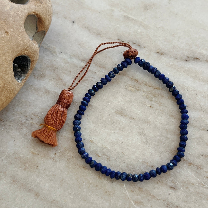 A faceted lapis bead bracelet with an adjustable tassel closure, displayed on a textured surface.