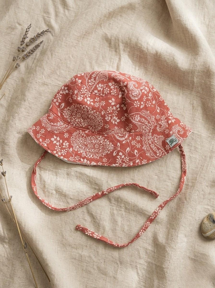 A flat lay of the Verano baby bucket hat in a warm terracotta orange, featuring a white paisley and floral pattern organic cotton muslin fabric. The hat includes matching chin ties and a small green turtle brand logo on the brim. It is styled on a neutral linen fabric with a sprig of dried lavender and a small smooth stone.