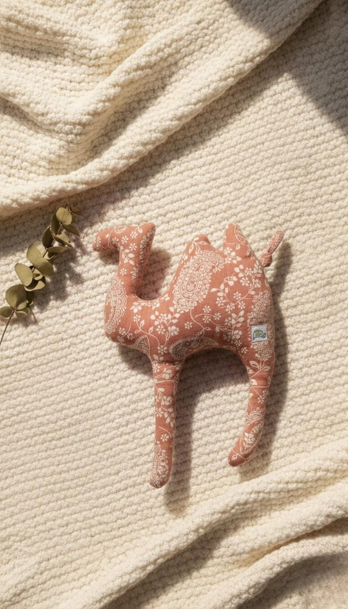 A top-down photograph of a plush cushion shaped like a camel, featuring a terracotta-colored fabric with an intricate white paisley and floral pattern. The camel cushion is styled on a textured, cream-colored knitted blanket in soft sunlight, with a small sprig of dried green eucalyptus leaves to the left.