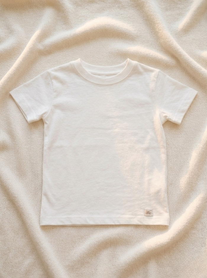 Handmade white baby T-shirt in soft breathable cotton, minimalist short-sleeve design by La Tortuga Marisa, laid on neutral textured fabric background