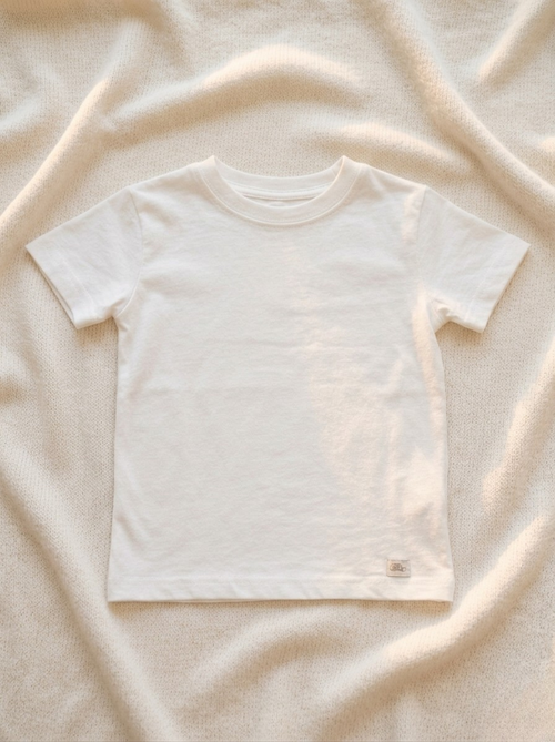 Handmade white baby T-shirt in soft breathable cotton, minimalist short-sleeve design by La Tortuga Marisa, laid on neutral textured fabric background