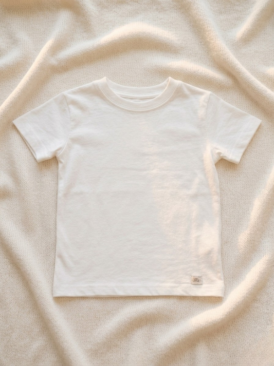 Handmade white baby T-shirt in soft breathable cotton, minimalist short-sleeve design by La Tortuga Marisa, laid on neutral textured fabric background