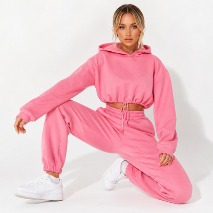 Women tracksuit set in pink on a female model with cropped hoodie, matching joggers and sneakers on a clean white backdrop.
