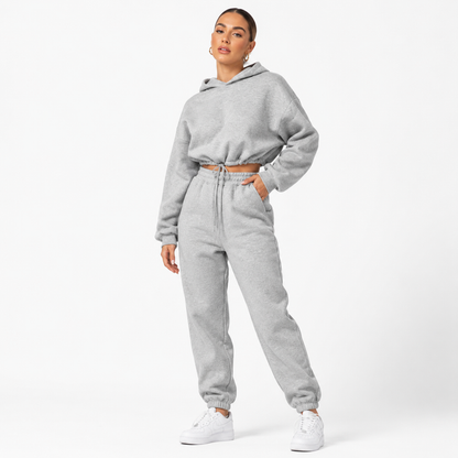Women tracksuit set in light gray on a female model with cropped hoodie, matching joggers and sneakers on a white studio backdrop.