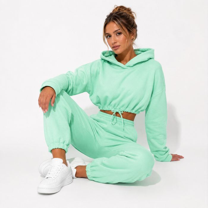 Women's green hoodie and jogger set shown on a female model in a seated studio pose against a clean white background.