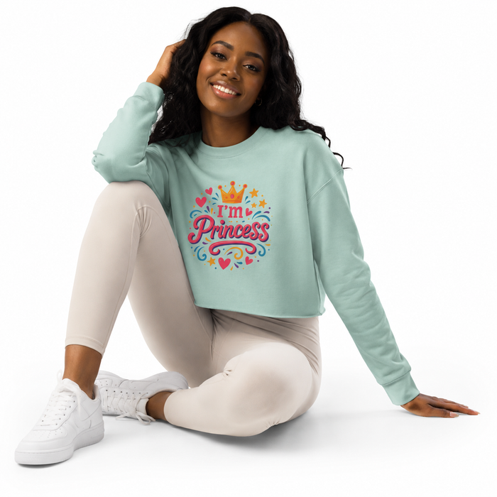 A woman sitting on a white studio floor wearing a mint green cropped sweatshirt with a colorful 'I'm Princess' graphic, paired with light beige leggings and white sneakers.